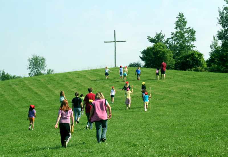 people walking on grass to a cross on the hill ahead of them